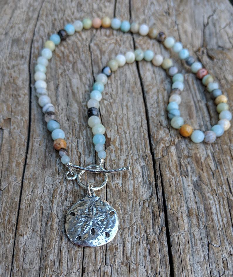 This handmade artisan one-of-a-kind amazonite necklace features the beautiful amazonite gemstone complimented by sterling silver. The calming pastel blues and greens of amazonite together with the silver sand dollar radiate peaceful energy and tranquility. Feel like you are taking a walk on a white sand beach with this beautiful unique necklace. 