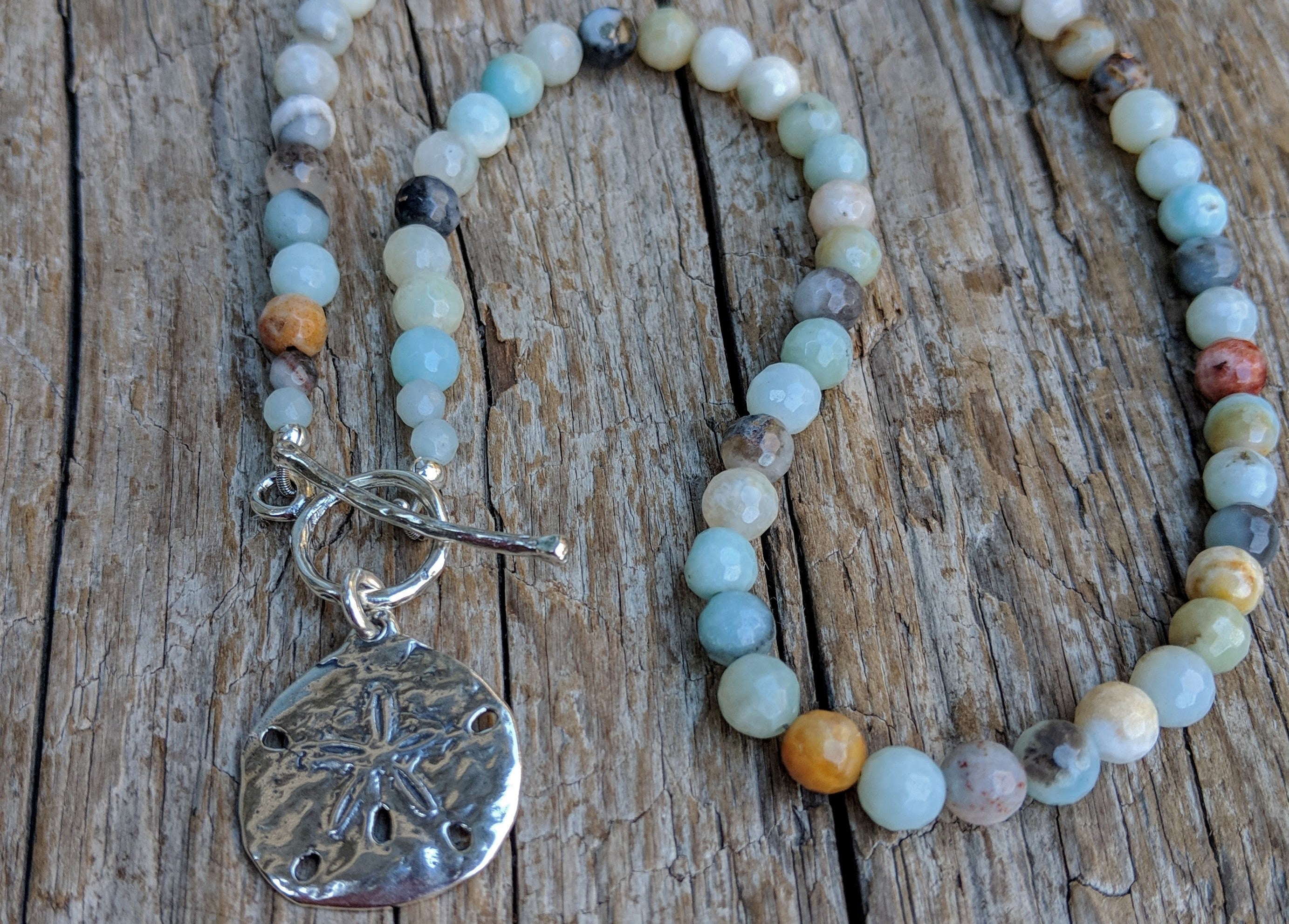 This handmade artisan one-of-a-kind amazonite necklace features the beautiful amazonite gemstone complimented by sterling silver. The calming pastel blues and greens of amazonite together with the silver sand dollar radiate peaceful energy and tranquility. Feel like you are taking a walk on a white sand beach with this beautiful unique necklace. 