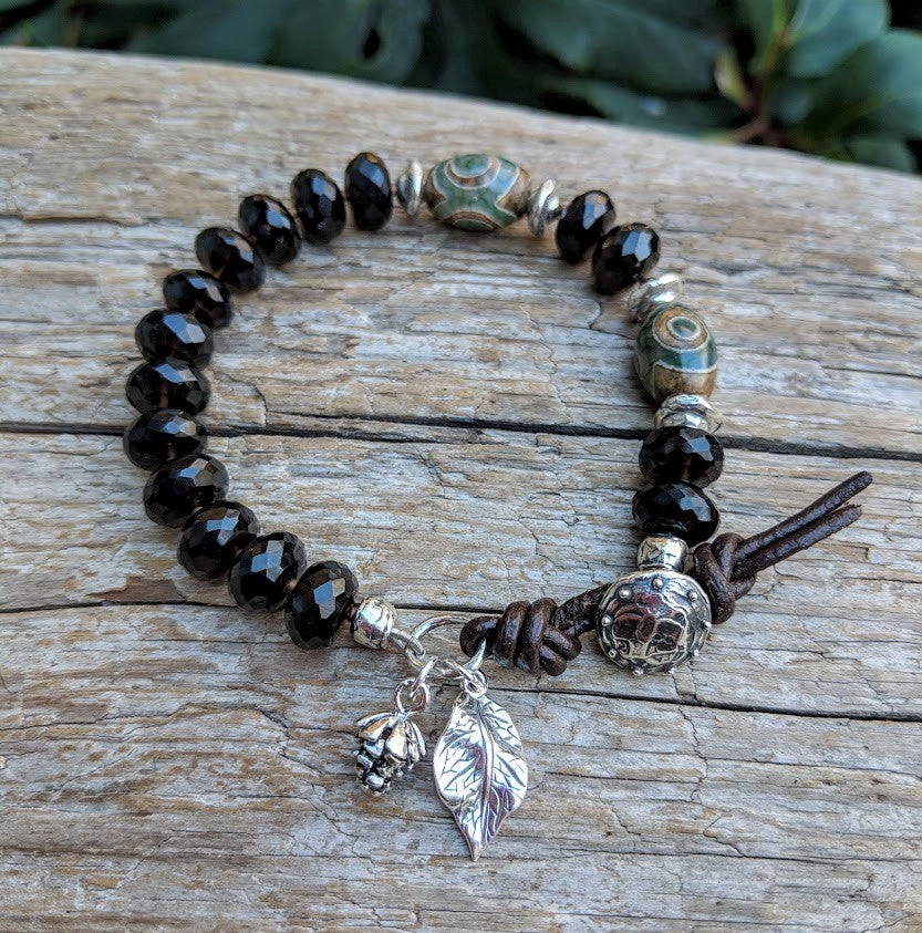 A gorgeous and bold handmade artisan bracelet showcasing the beautiful natural dark brown smoky quartz and green agate complimented by sterling silver pine cone and leaf charms and leather accents. Wearing this bracelet feels like taking a walk through the woods, with the sterling silver leaf and pine cone charms creating a forest theme. 
