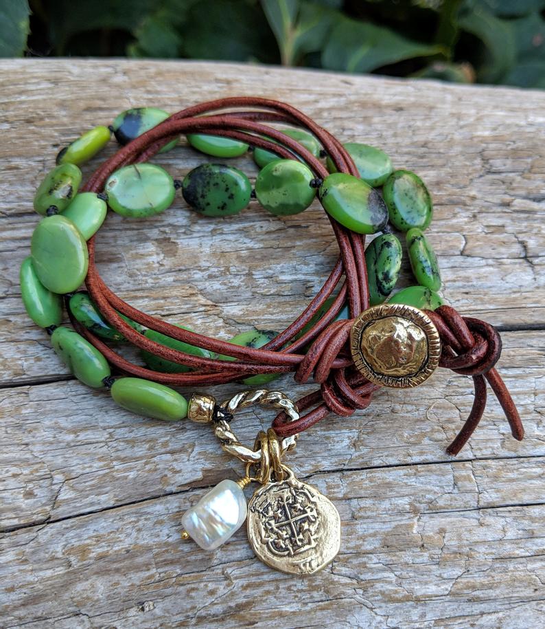 This handmade artisan bracelet combines the gorgeous forest greens of chrysoprase with natural leather, pearl, and gold bronze elements. The brown leather adds a contrasting texture to the combination, while complementing the colors of the stone. The Spanish cross Reale coin charm creates a rustic-medieval theme. A white pearl accent adds a fresh contrast to the combination of natural greens. 