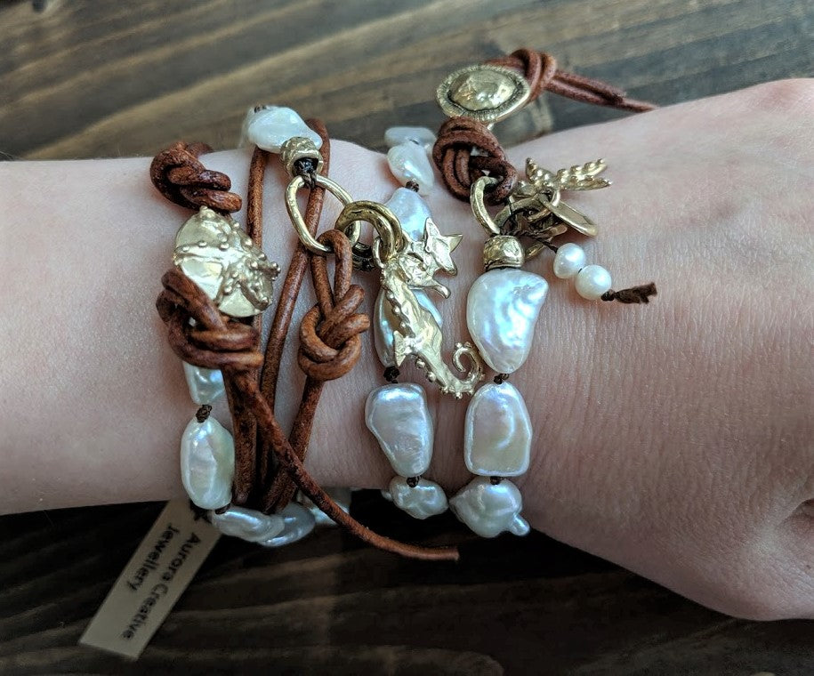This gorgeous handmade artisan wrap bracelet-necklace combines the beautiful large white baroque pearls with gold bronze and leather elements. The gold bronze button, seahorse and star charms add a beautiful shine to the combination and create an ocean theme. The bracelet is held together by a silk thread and a brown leather cord. Wearing this bracelet feels like taking a night walk on the beach with the sky full of stars. 