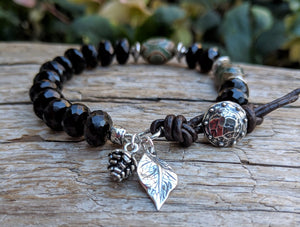 A gorgeous and bold handmade artisan bracelet showcasing the beautiful natural dark brown smoky quartz and green agate complimented by sterling silver pine cone and leaf charms and leather accents. Wearing this bracelet feels like taking a walk through the woods, with the sterling silver leaf and pine cone charms creating a forest theme. 